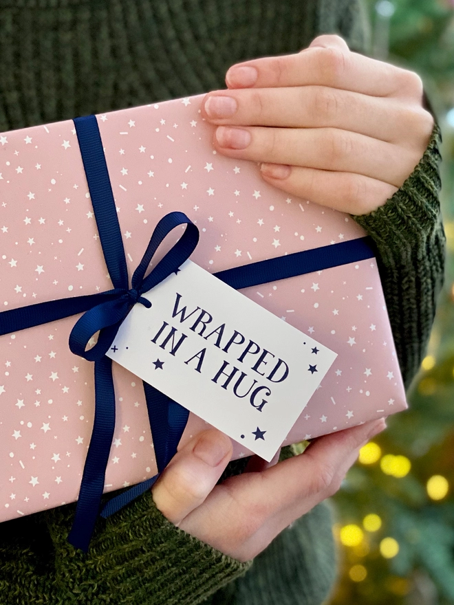 A gift wrapped in a gentle pink start design wrapping paper, tied with navy blue ribbon, with a gift tag that reads "Wrapped in a hug" is being held in front of a Christmas Tree.