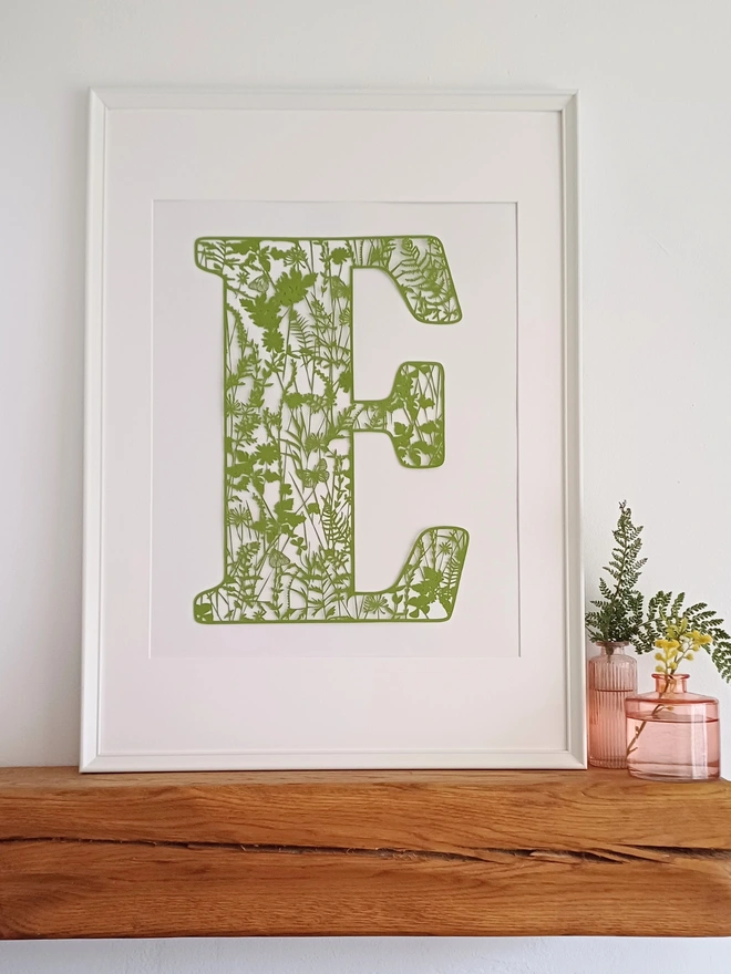 Letter E botannical papercut featuring summer flowers and plants in olive green displayed in a white frame resting on a wooden mantelpiece