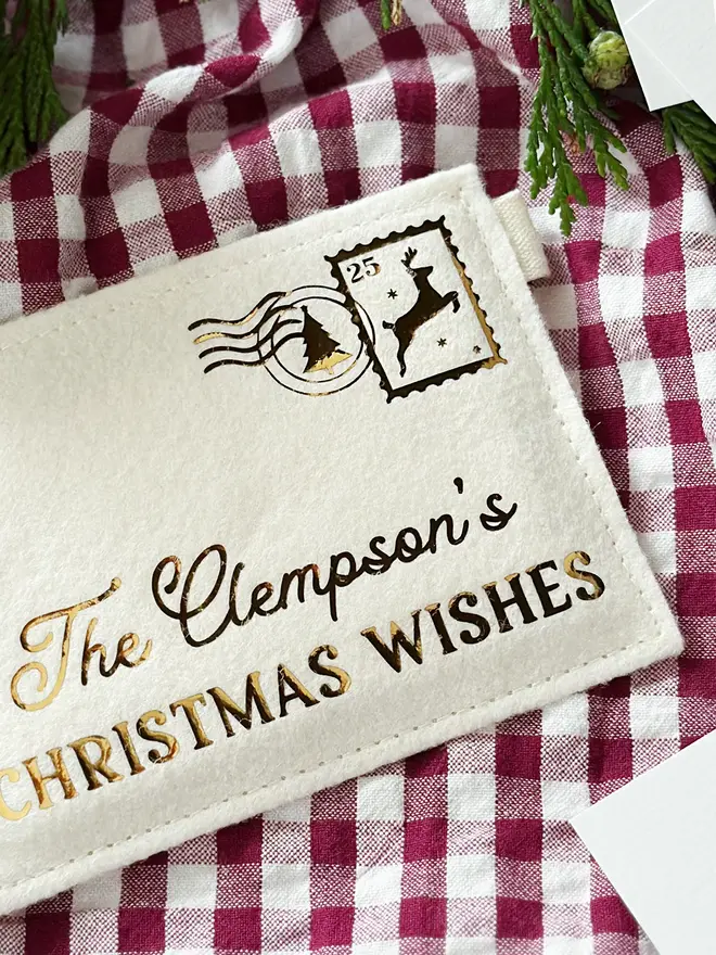 A cream felt personalised envelope reading "The Clempson's Christmas Wishes," and featuring a golden reindeer postage stamp is placed on a red gingham cloth.
