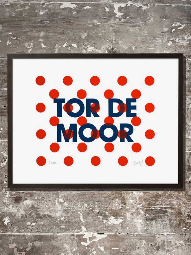  A flat lay view of a framed print, Tor de Moor words screenprinted in dark blue, with red polka dots printed in red on top. All lying on a wooden paint flecked floor.