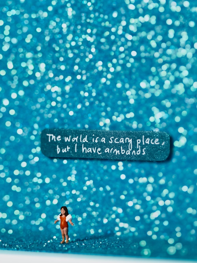 Miniature scene in an artbox showing a tiny girl in armbands against a sparkling turquoise glittery backdrop ,evoking a swimming pool. The words “The world is a scary place but I have armbands” are written on the back wall.