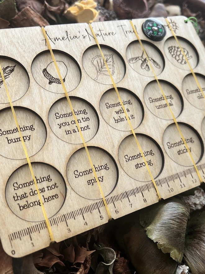 A personalised wooden nature scavenger hunt board displayed on an outdoor surface. The board is rectangular with rounded corners and engraved with 15 small collection slots held by clear bands. A custom name is engraved at the top, and a button compass with a wrist lanyard is attached for carrying. The wood has a smooth, light finish, and no items are placed in the slots; only the board is shown.