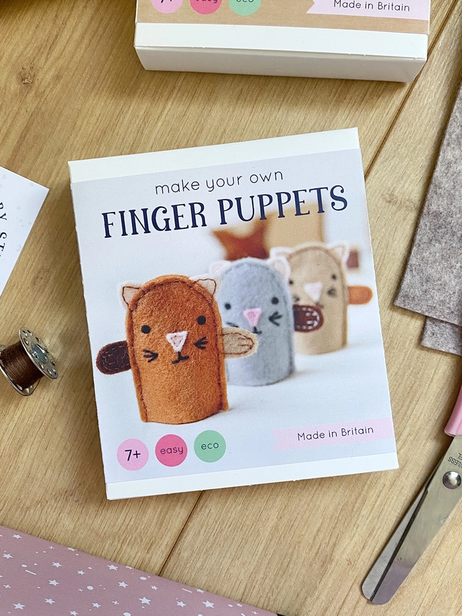 A craft kit to make three felt kitten finger puppets lays on a wooden desk beside various materials and scissors.