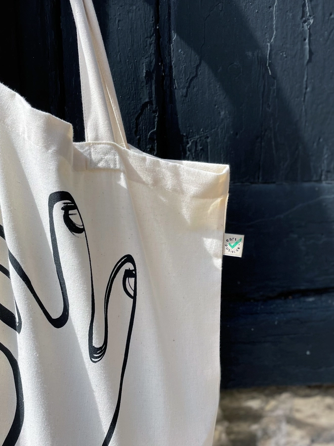 Close up of Ok tote bag hanging on old dark blue/grey doors. Showing some of the ink printed and the fairtrade label.