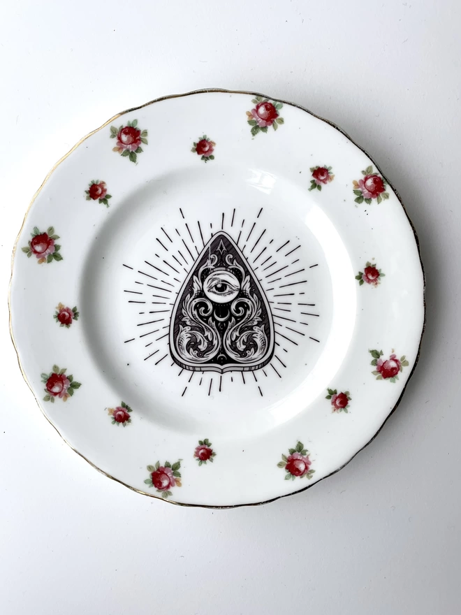 vintage plate with an ornate border, with a printed vintage illustration of a Ouija Planchette in the middle 