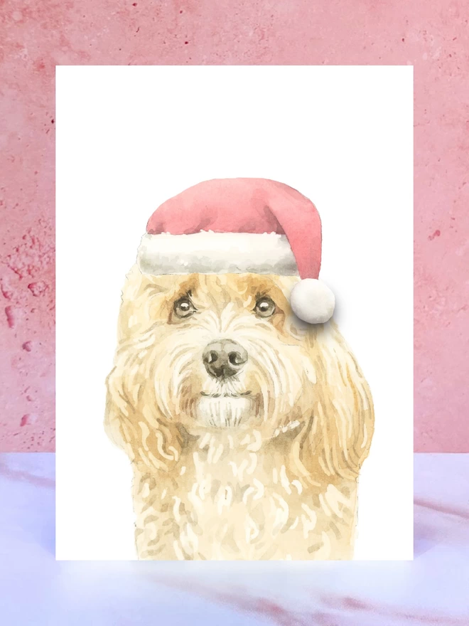 A Christmas card featuring a hand painted design of an apricot cavapoo, stood upright on a marble surface surrounded by pompoms. 
