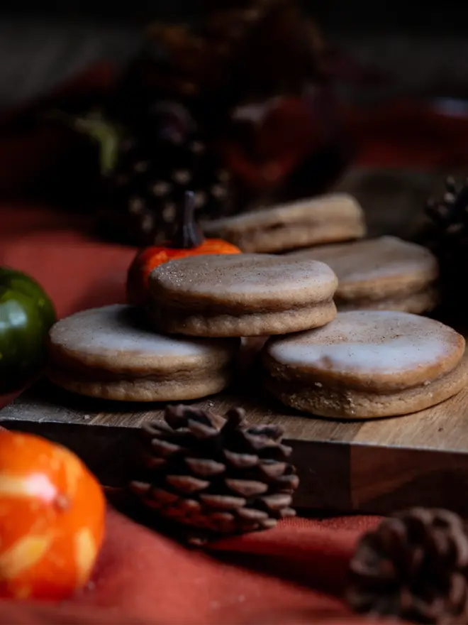 Pumpkin Spice Welsh Cakes on a wooden chopping board, an orange table cloth, and surrounded by autumnal decorations such as pumpkins and pine cones.
