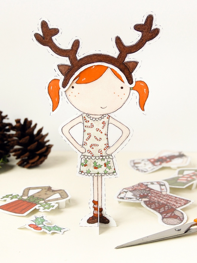A Christmas paper doll with cut out outfits, from wrapping paper, stands on a white table.