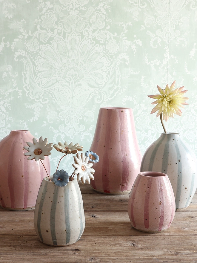 Handmade Ceramic Stoneware Rounded Vase in Blue and White Stripe holding a dark red flower stem. The vase sits on a teal hardback book with a collection of three pink sea urchins sat beside it on a wooden surface. 