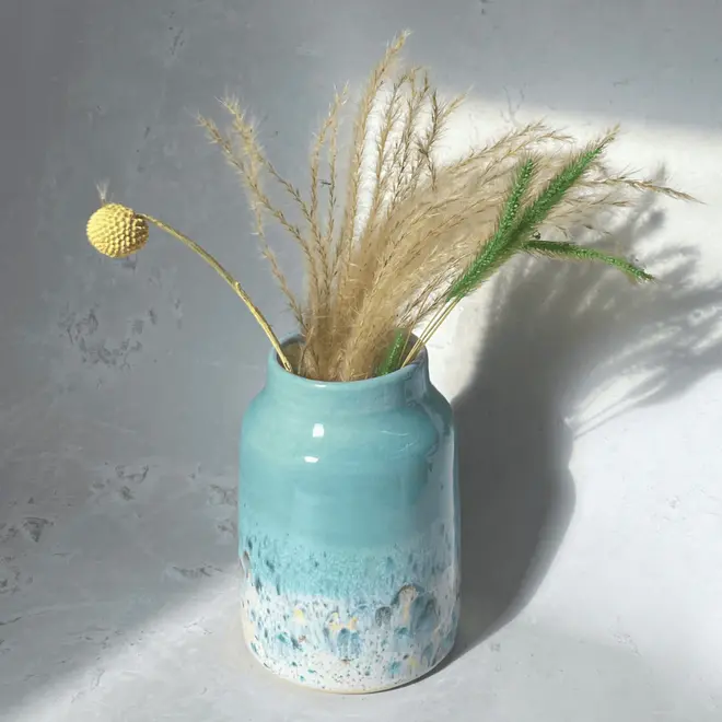Shoreline Large Vase, a colourful vase placed on a stone surface in front of a plain backdrop. It is accompanied by flowers. 