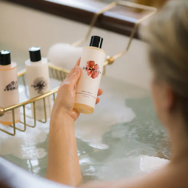 person off-camera, in a bath tub, holding a white bottle of body wash with pink logo and black cap, above a tub of bubbly water