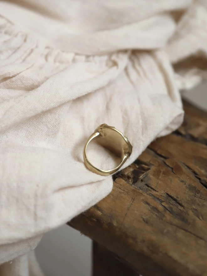 An image of the back and underside of a hand carved gold toned brass sun ring, resting on cream coloured linen type fabric and rustic wood 
