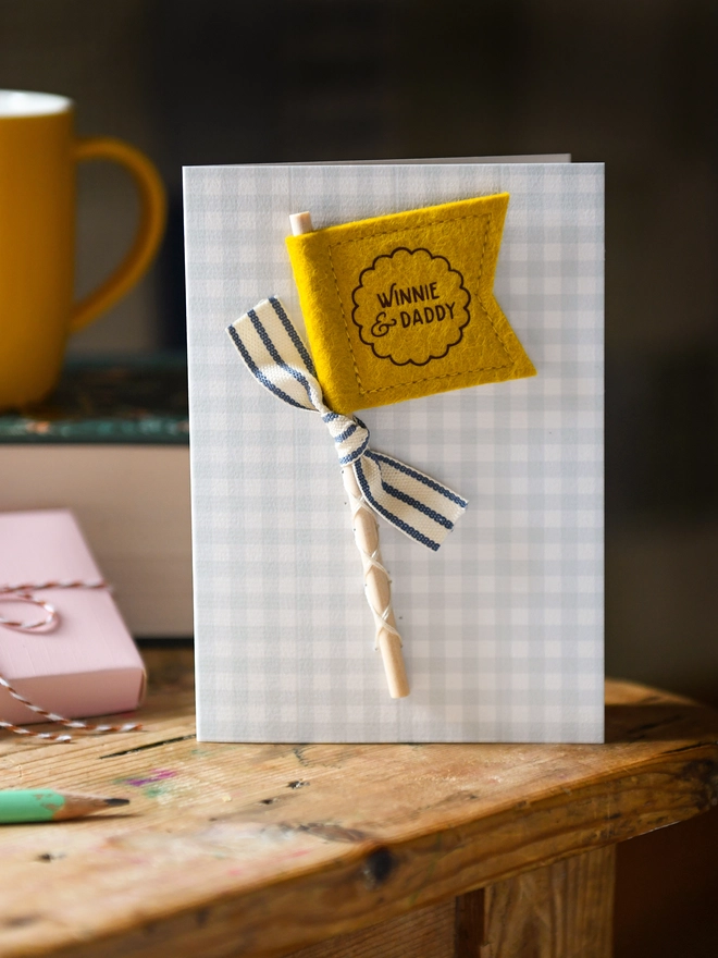 Personalised Father’s Day card with mustard felt flag reading “Henry & Daddy”, handmade keepsake card with ribbon and wooden stick detail. The card stands on a wooden table beside a book, mug, candle, and wrapped gift.