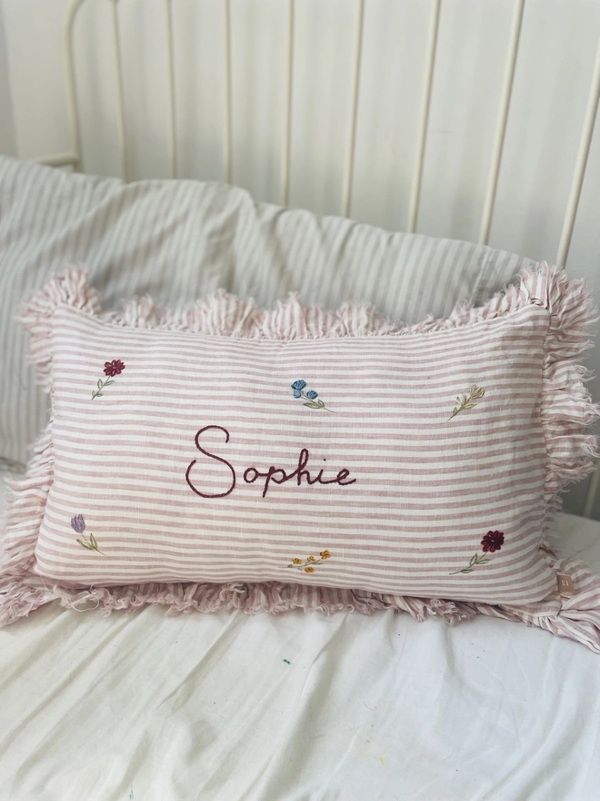     Personalised pink striped stonewashed linen cushion with ruffle trim, hand-painted ditsy florals and hand-embroidered name, styled on a child’s bed.