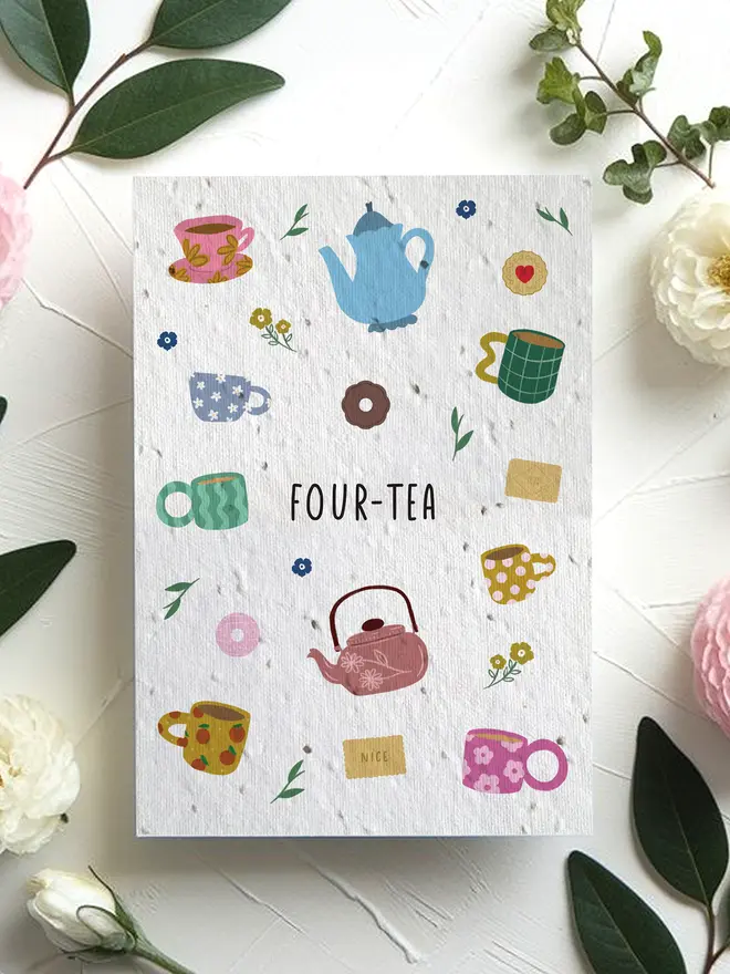 A greeting card titled “FOUR-TEA” featuring colorful illustrations of teapots, mugs, tea bags, and small flowers, surrounded by pink and white blossoms with green leaves on a white textured surface.