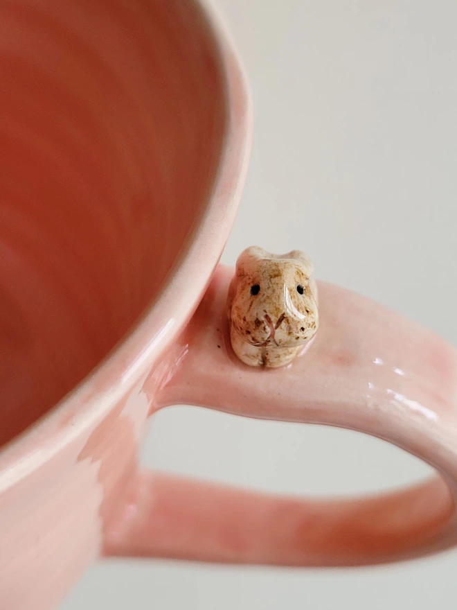 close up of a pink ceramic cup showing a little beige pottery bunny rabbit with a cute face on the top of the handle 