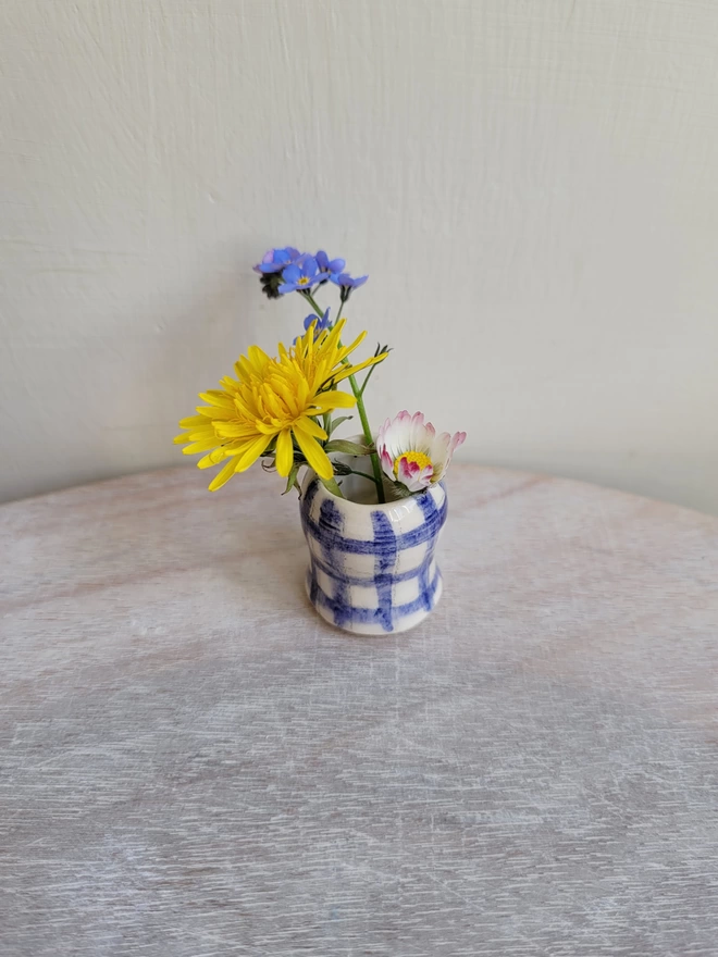 a tiny pottery blue gingham vase with forget me not  daisy and dandelion flower in 
