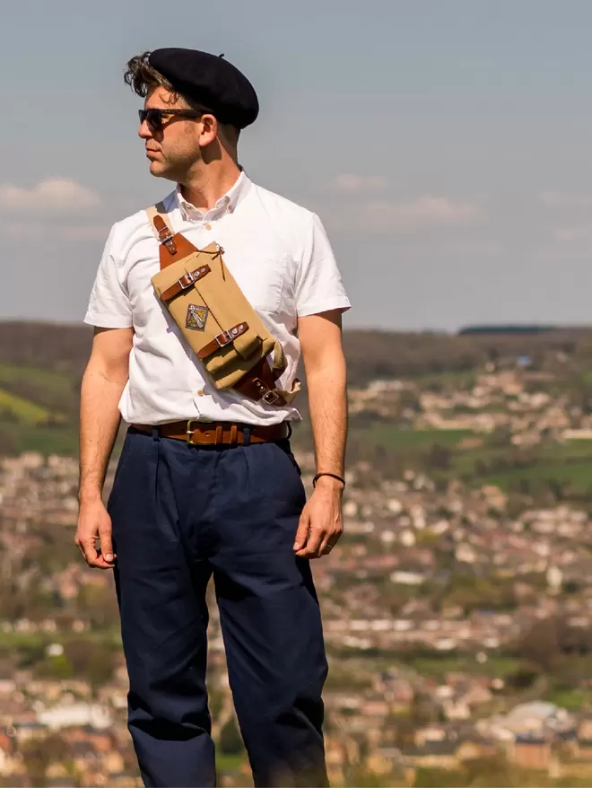 Model wearing Taupe Owlpen sling bag, white shirt and blue trousers with a village in the distant background.