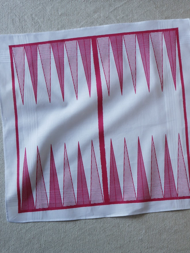 A Mr.PS Backgammon board game hankie printed in pink laid flat on a linen tablecloth
