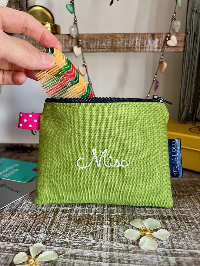 Small green zipped pouch with 'Misc' embroidered in white thread. A hand is placing a colourful comb inside it.