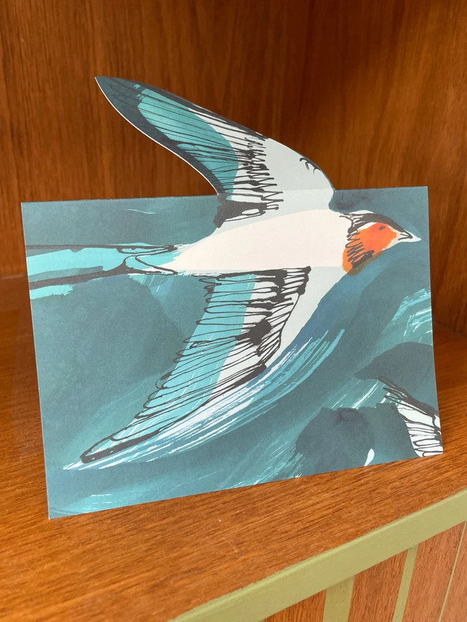 Esther Kent illustrated greeting card showing inky blue swallow illustration on a teal background. The swallow's wing pops u.