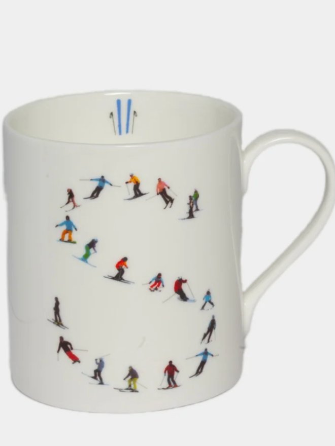 A simple white mug illustrated with an ’S’ made of small, multicoloured skiers. The mug is set against a plain, white background.