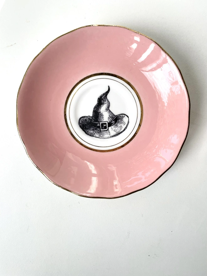 A blush pink plate with gold trim and a white centre, in the middle is a vintage black and white illustration of a witch's hat