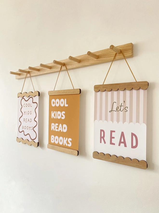 A wooden magnetic picture frame, strung with vegan suede. Hung on a wavy wooden wall dot. Displaying a colourful stripy kids print that says cool kids read books 