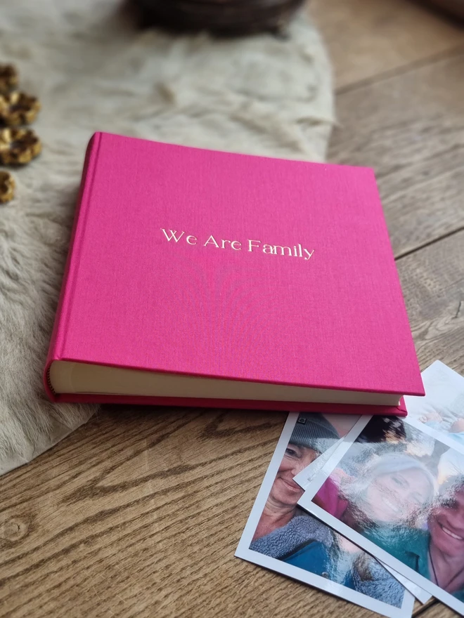 A hot pink photo album printed with 'We Are Family' in gold lettering sits on a wooden surface, next to a pile of photos.