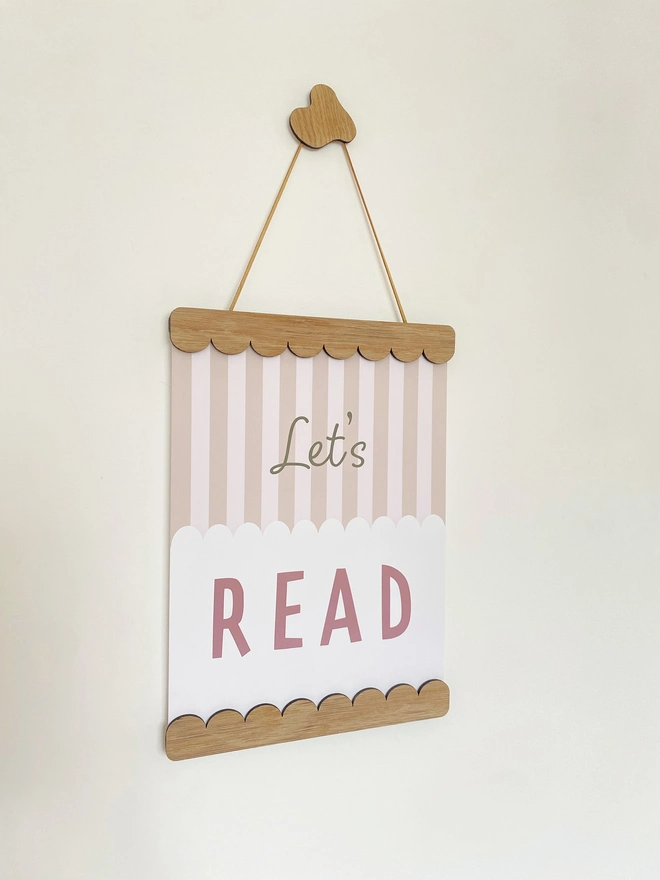 A wooden magnetic picture frame, strung with vegan suede. Hung on a wavy wooden wall dot. Displaying a colourful stripy kids print that says cool kids read books 
