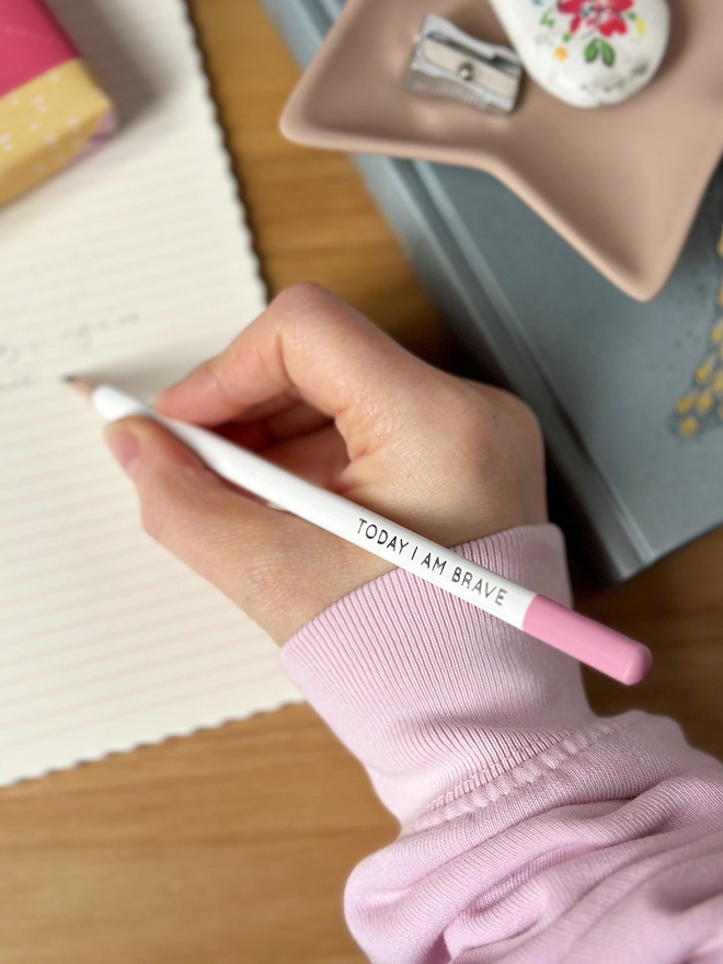A white pencil with a pink top and the words "Today I Am Brave" printed along the side is being held above an open notebook.