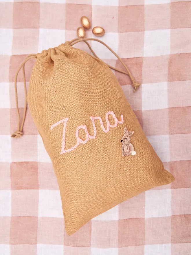 jute drawstring sack with brown knitted bunny decoration and pink 'zara' name next to golden chocolate eggs on red gingham table cloth
