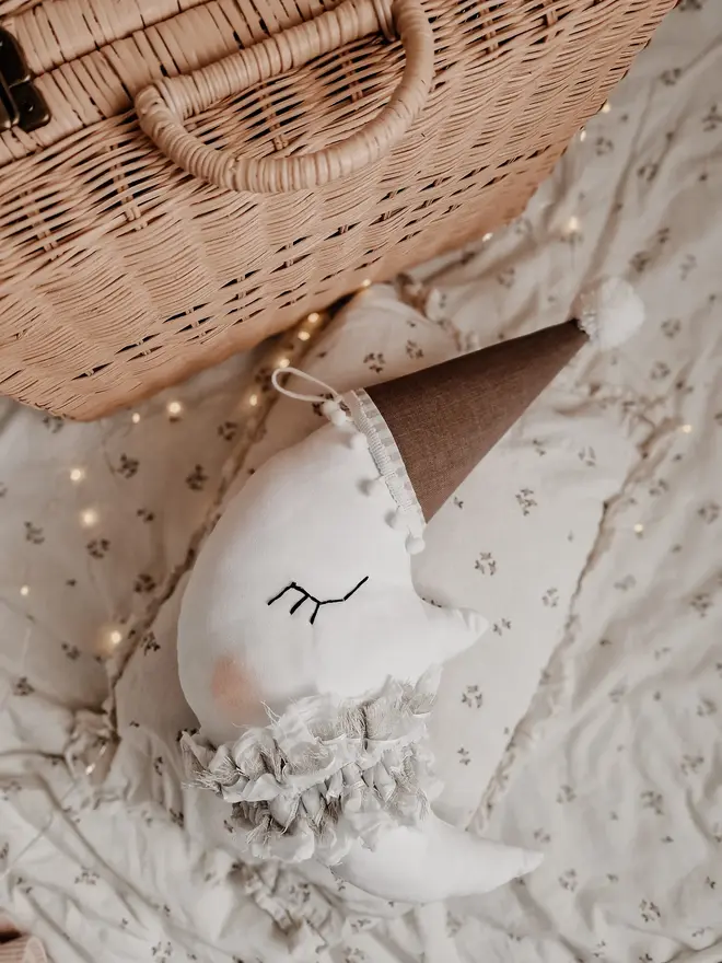 Hanging Moon Pillow Decoration, a hanging moon pillow decoration sitting against a wooden hamper accompanied by other bedding accessories. 