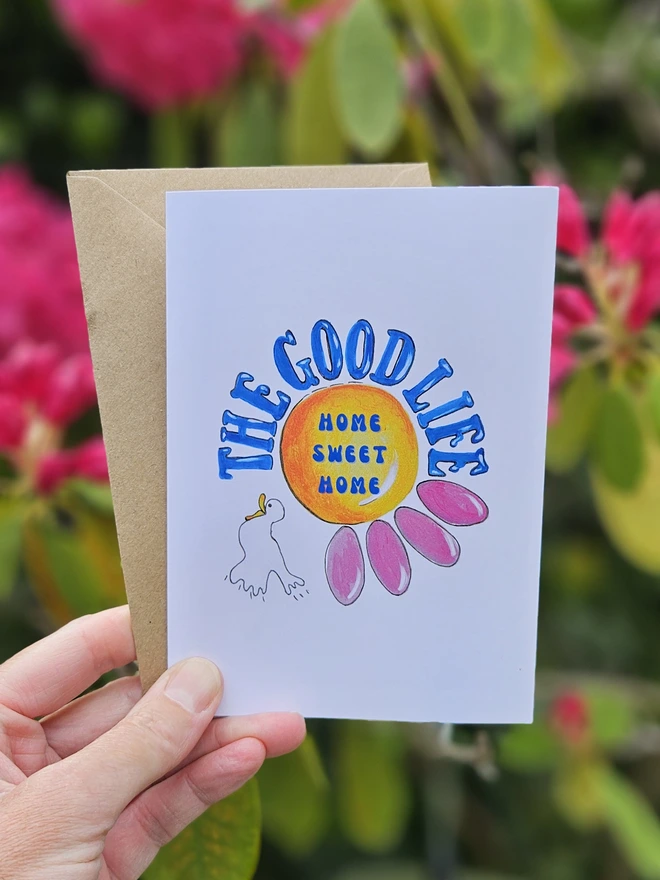 A white card with an illustration of a pink flower and a white bird. The Good Life is written using a retro font style in blue. In the centre of the flower it says Home Sweet Home. 