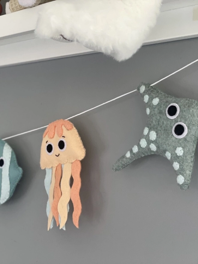 Nursery Under The Sea Garland, a sea themed garland hanging from a white shelf against a grey backdrop in a child's bedroom. 