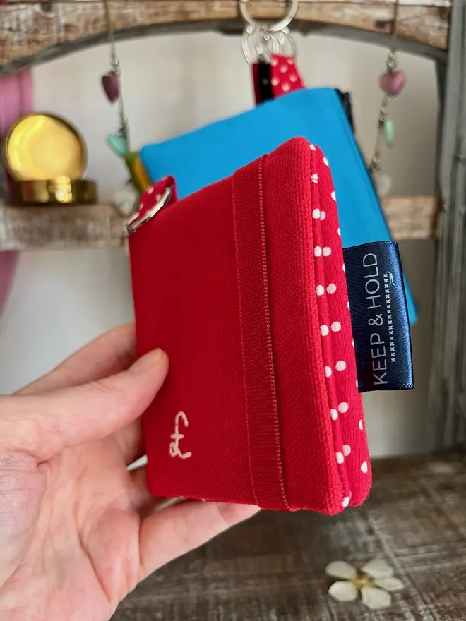 Small red card wallet with key ring, a hand holding it to show the side view with polka dot lining peeking through.