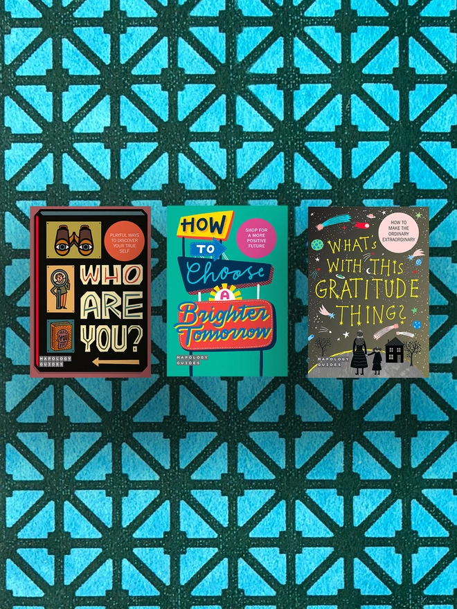 A bundle of three front Mapology Guides covers on a multi-coloured and multi-patterned background. The maps are: Cover of Mapology Guide titled “Who Are You?” featuring retro-style illustrations of binoculars, a person holding a magnifying glass, and a book titled “Understanding You.” Tagline reads “Playful ways to discover your true self.” Cover of Mapology Guide titled “How to Choose a Brighter Tomorrow” featuring bright retro signage and playful typography promoting optimism and hope. Cover of Mapology Guide titled “What’s With This Gratitude Thing?” featuring a starry night sky, planets, comets and two figures standing near a house. Text reads “How to make the ordinary extraordinary.”
