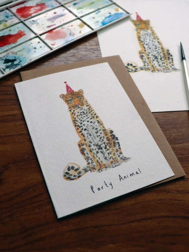 Alternative View Of The Leopard Party Animal Birthday Card Sitting Slightly Out Of Shot Is The Original Hand Painted Illustration and, Paint Pallet and Paint Brush 