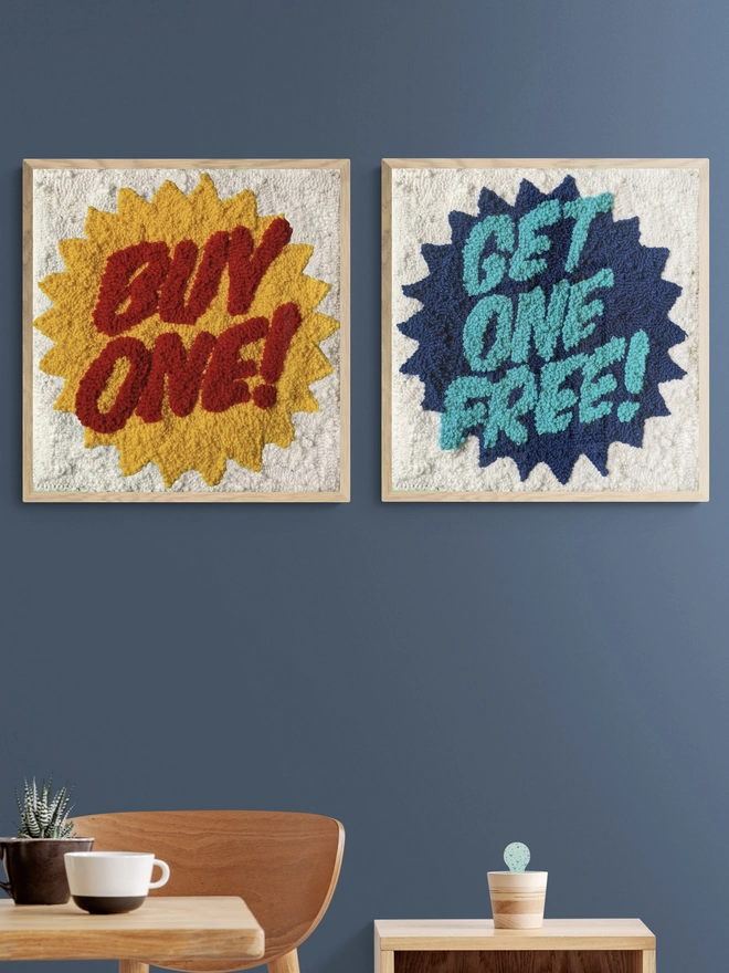 Two textile are pieces in square wood frames read "buy one", "get one free" mounted on a blue wall behind a wooden table and chairs