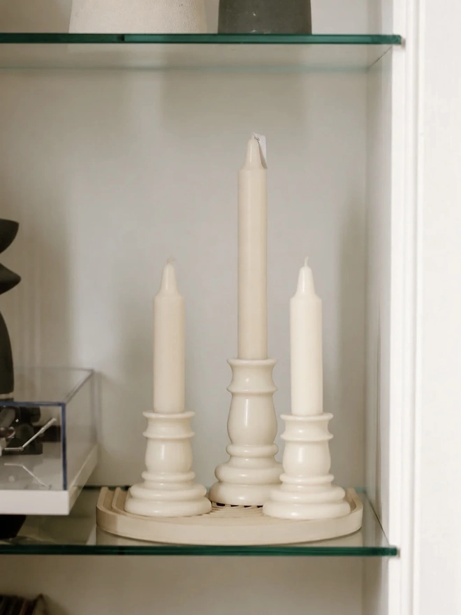 Baluster Christmas Candles (Set Of 2). Three unlit cream candlestick candles in tall and short heights, grouped on a ribbed stone tray on a glass shelf. The coordinating trio makes an elegant display for shelves, mantelpieces and sideboards.
