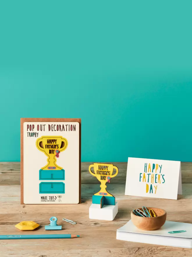 3D Happy Father's Day trophy laser-cut decoration and Happy Father's Day card and brown kraft envelope on top of a wooden desk in front of a teal coloured background