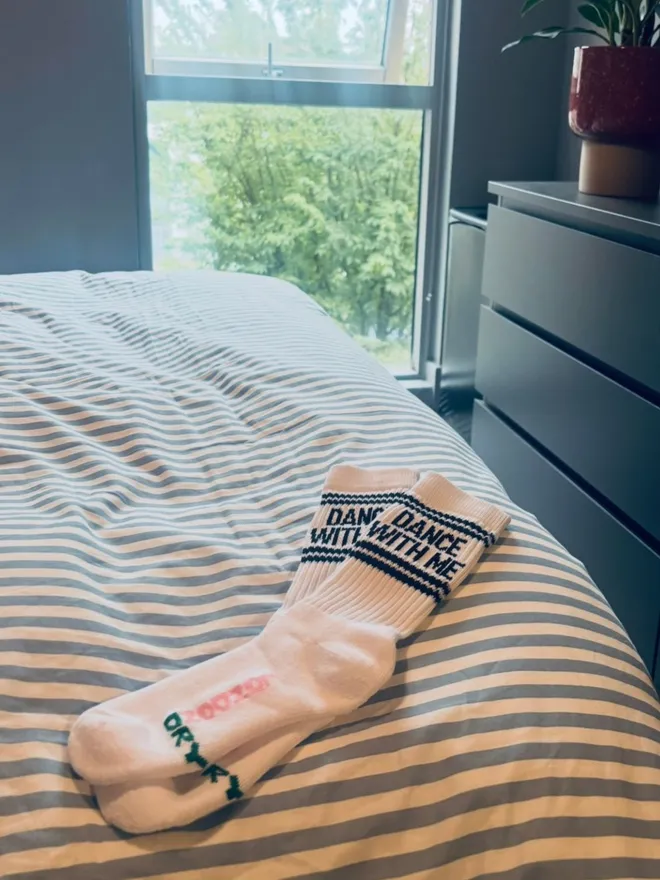Socks with black DANCE WITH ME text on them laying on a bed with window and chest of drawers in background