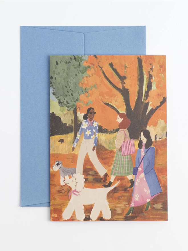 A greetings card featuring three women walking three dogs in a beautiful autumnal parkland. The women are smiling and chatting as they walk. In the background there is long grass and a brown labrador can be seen poking their head out excitedly. 