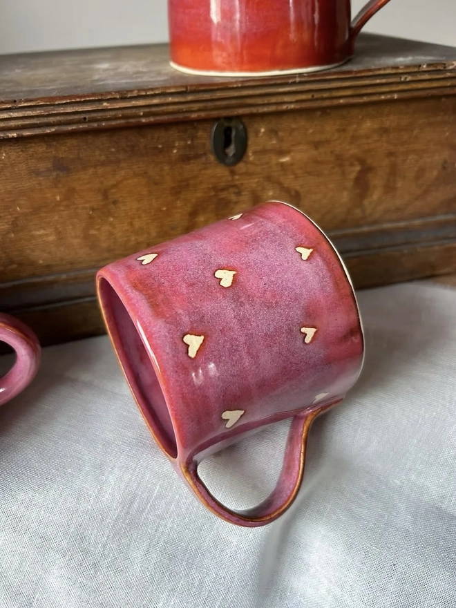 Love Heart Mug Raspberry, a red heart mug on a white surface in front of another mug on a wooden surface in the background. 