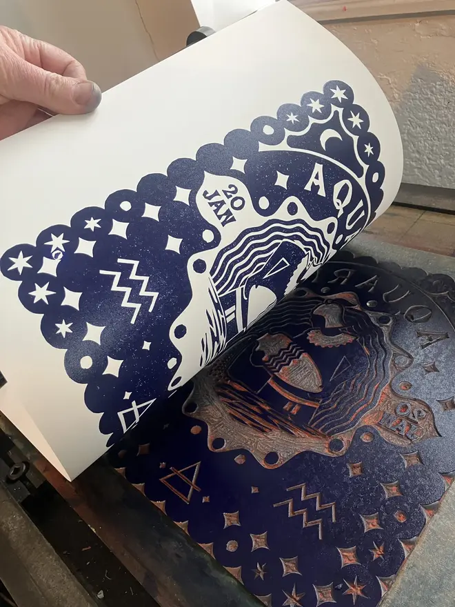 aquarius zodiac linocut. a linocut image of a man pouring water and zodiac symbols, printed on aa blue background.