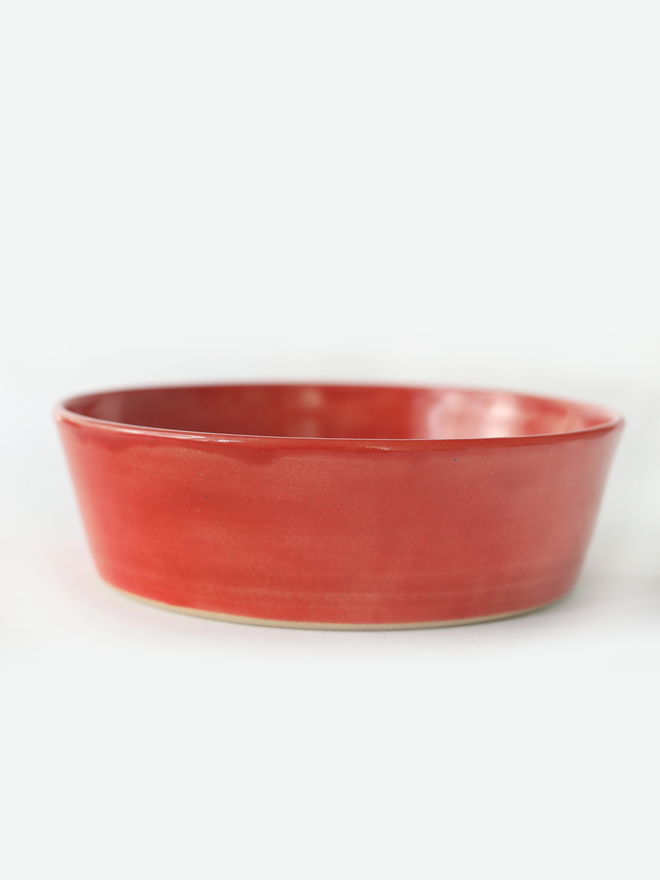 A single wide, shallow glazed ceramic pasta bowl in cherry red, photographed from a slight angle against a plain light grey background, showing the unglazed natural clay base.