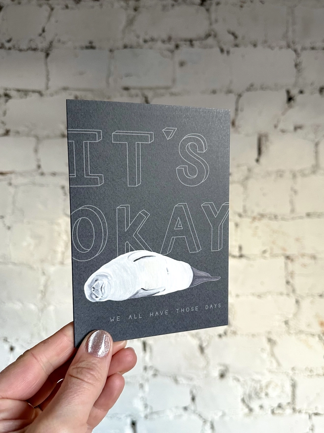 a dark grey greetings card featuring an illustration of a seal with the text “It’s Okay - we all have those days”