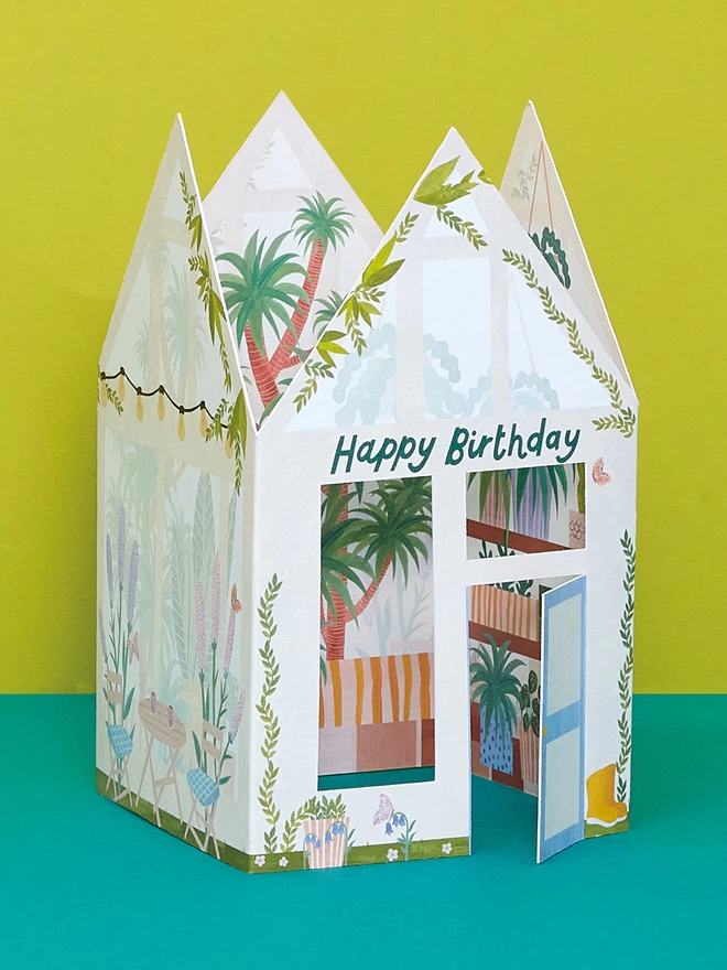 A beautiful 3D greenhouse card features hand painted plants and foilage with die cut windows and a door that opens to reveal the inside bursting full of tropical plants and colourful pots. A ‘Happy Birthday’ message in blue hand lettering sits at the top of the greenhouse