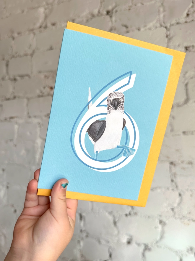 a bright blue card with a big white number 6 and an illustrated blue footed booby with a bright yellow envelope
