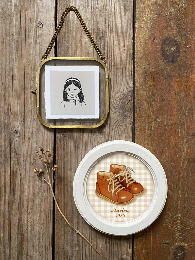 photograph of framed illustrated art work one frame is of a little girl drawing the 2nd is of a pair of brown baby boots with a gingham background one frame is round one is galss